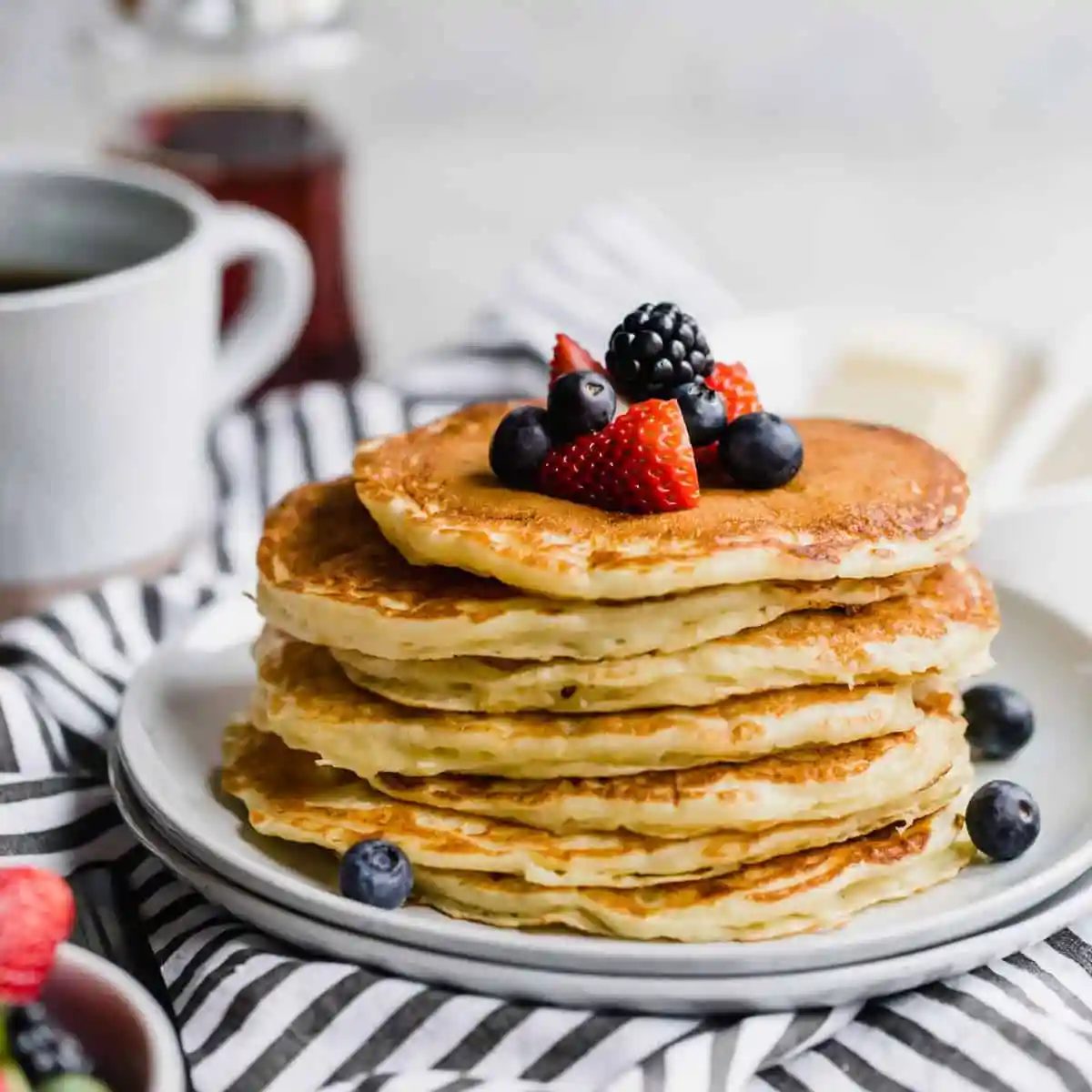 A stack of buttermilk pancakes topped with fresh berries on a white plate with a blue and white striped towel below.