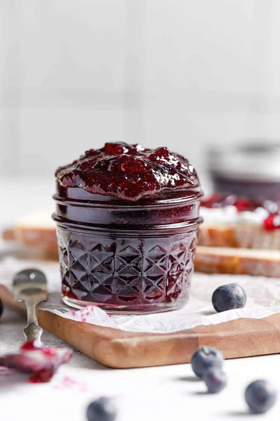 A wooden cutting board topped with parchment paper with a glass jar of blueberry jam and a silver spoon to the left.