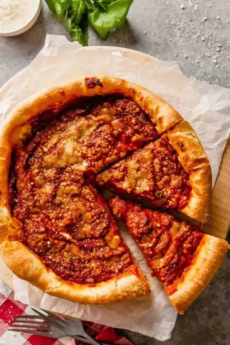 A deep dish pizza with a slice cut out on a slice of parchment paper with basil in the top left corner.