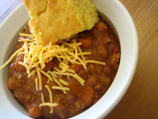 Chili in a white bowl with a piece of cornbread topped with shredded cheese.