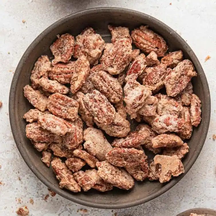 A square photo with candied pecans in a brown bowl on a white counter.