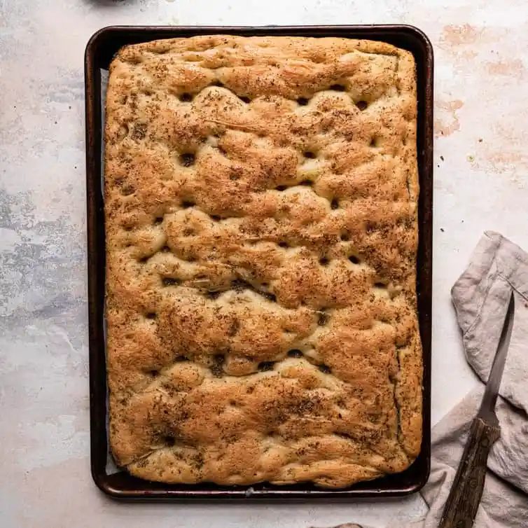 A top down photo of a baking sheet lined with parchment paper and fresh baked focaccia next to a bread knife on the right.