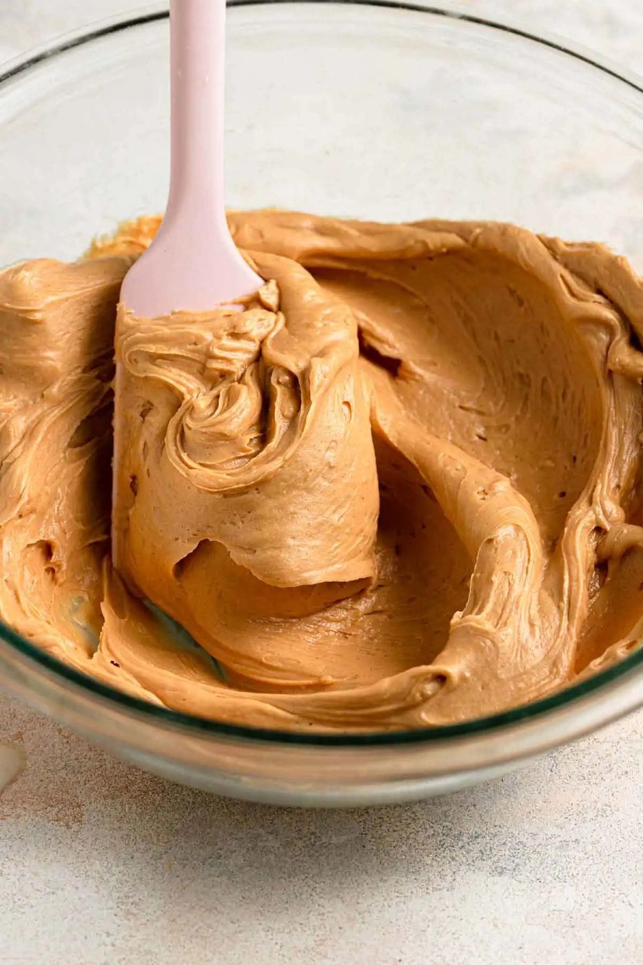 A pink spatula stirring peanut butter frosting in a glass bowl.
