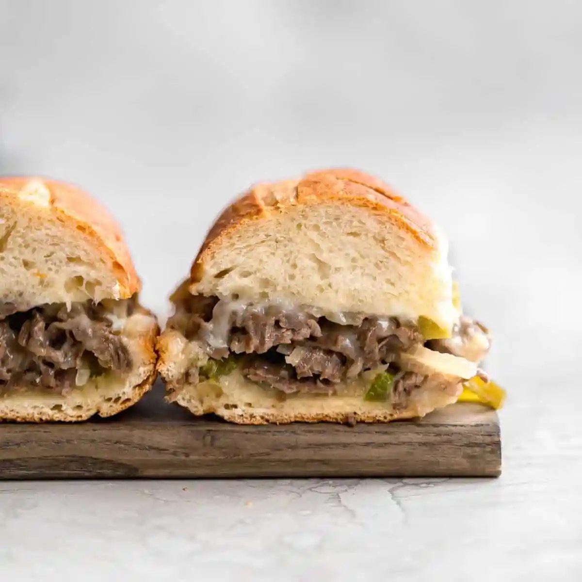 Two philly cheesesteak sandwiches on a wooden cutting board with the insides exposed showing the cheesy meat filling.