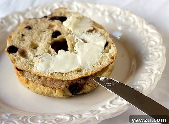A delectable blueberry bagel, expertly sliced in half and generously smeared with creamy cream cheese, awaiting enjoyment on a white plate alongside a butter knife.