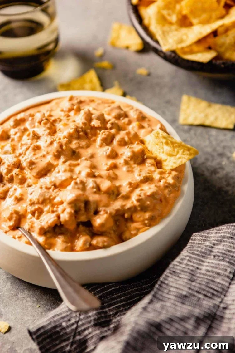 A white bowl of sausage dip on a grey counter with a spoon in the left side of the bowl and tortilla chips on the back of the counter.