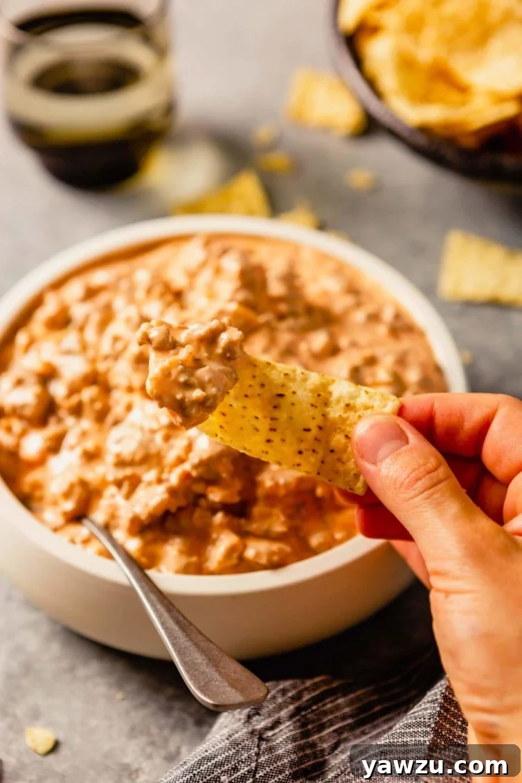 A white bowl with sausage dip and a hand holding a chip in front after dipping in the sausage dip.