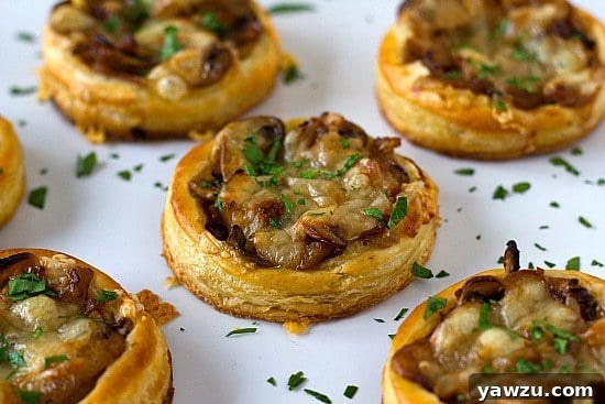 Golden brown caramelized onion, mushroom, and gruyere tartlets on a white serving plate, garnished with fresh herbs.