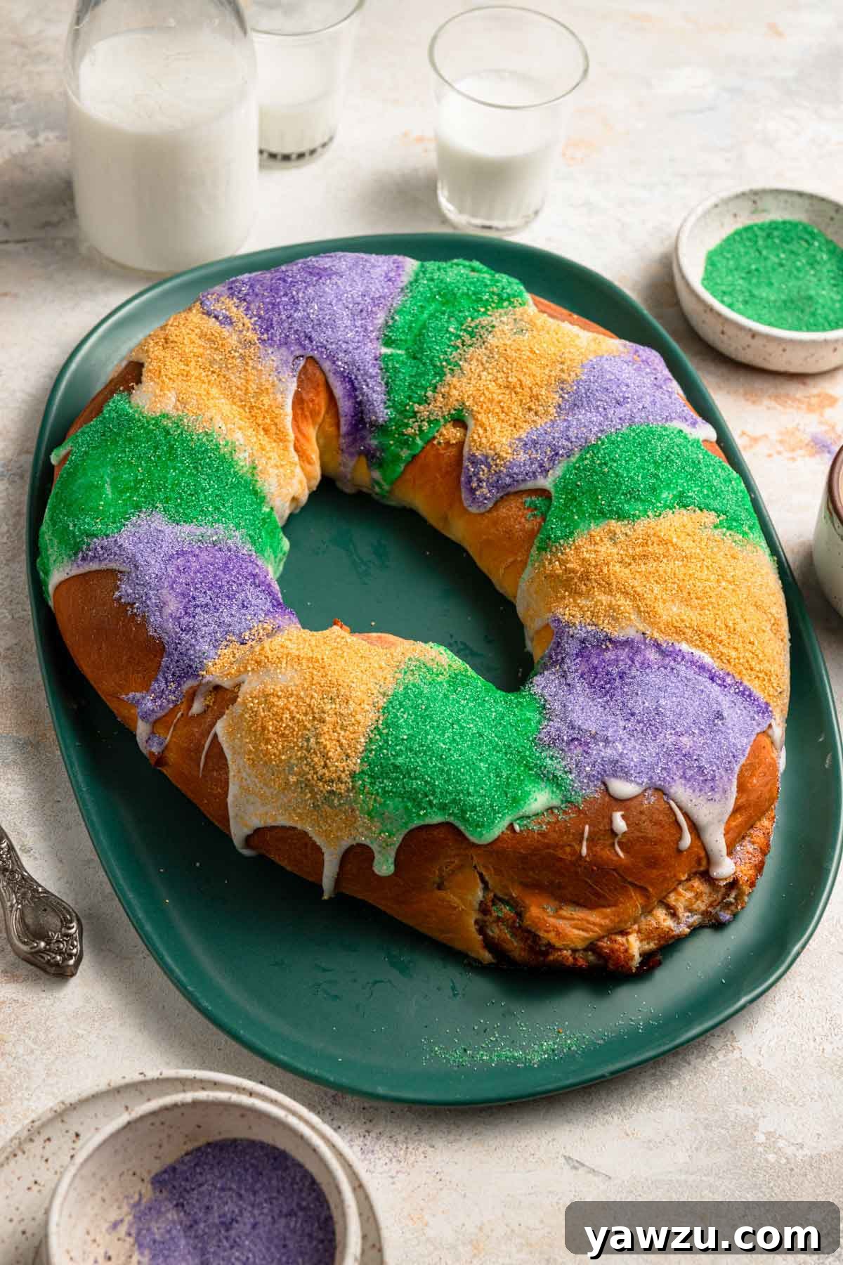 Photo of baked king cake on a green platter iced and decorated with sanding sugar in alternating strips of purple, green, and yellow.