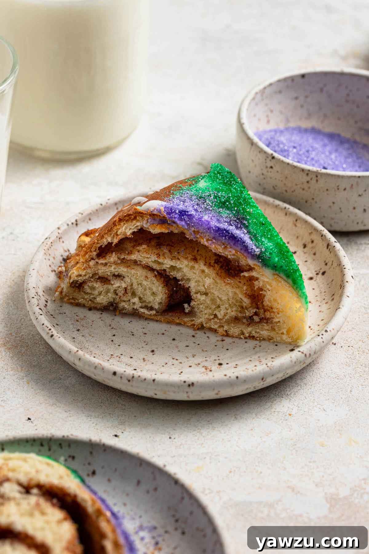 A slice of king cake on a speckled plate with a cup of milk and bowls of colored sanding sugar in the background.