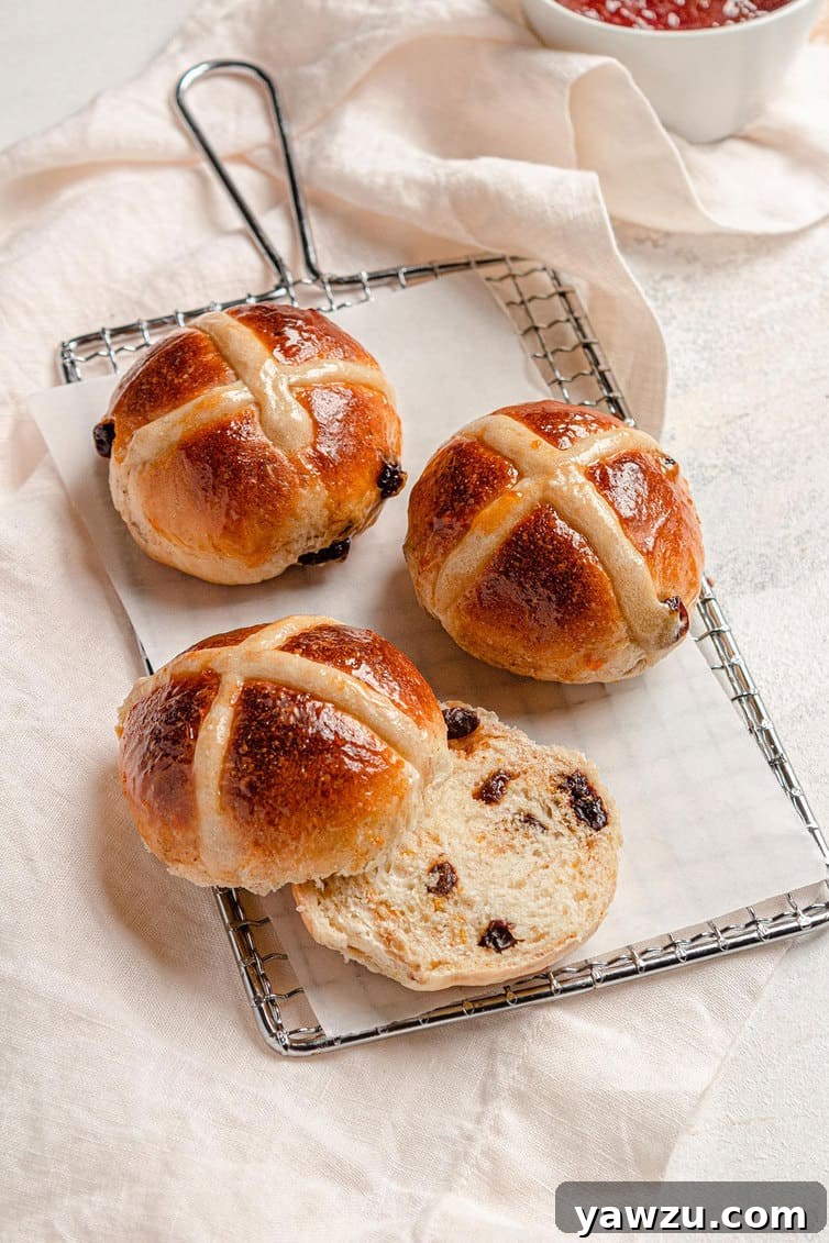 Three hot cross buns on a serving plate, one sliced in half, revealing its fluffy interior.