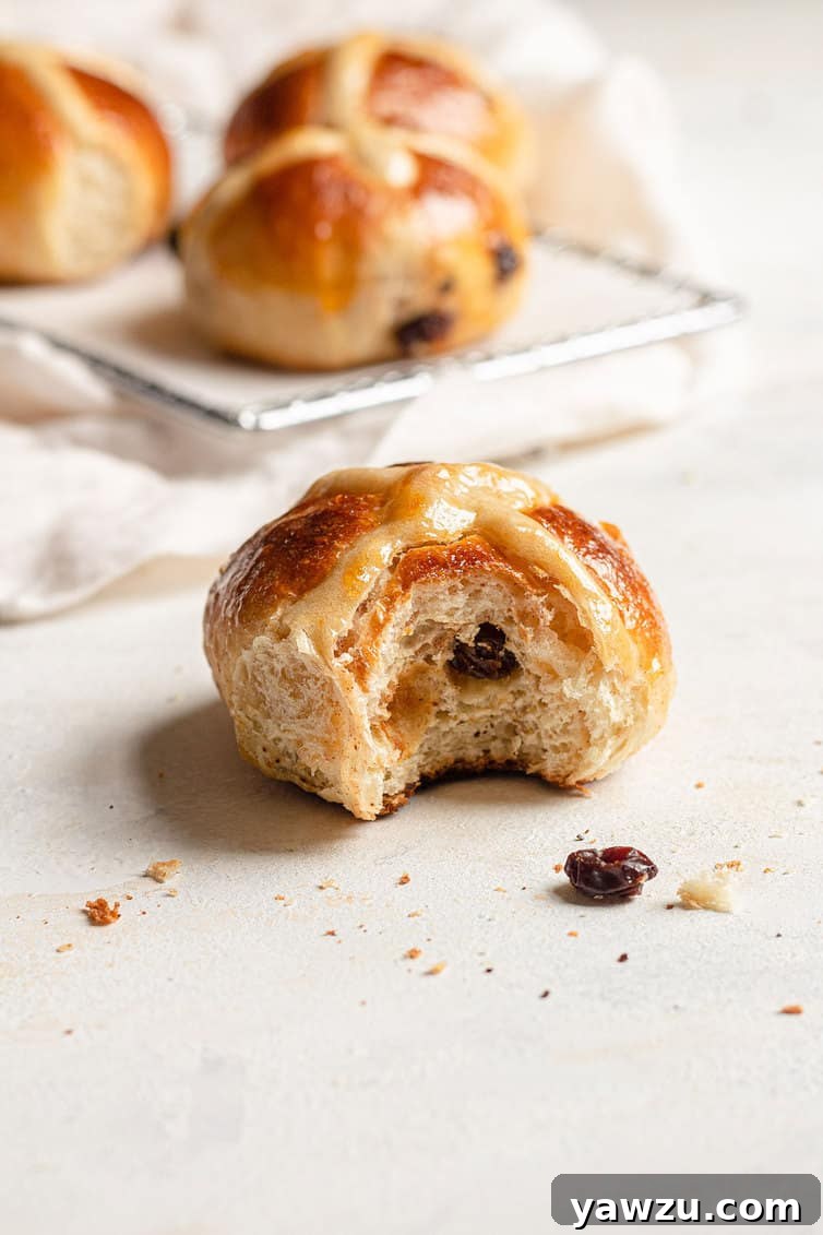 A hot cross bun with a bite taken out, revealing the soft interior and raisins, with other buns blurred in the background.