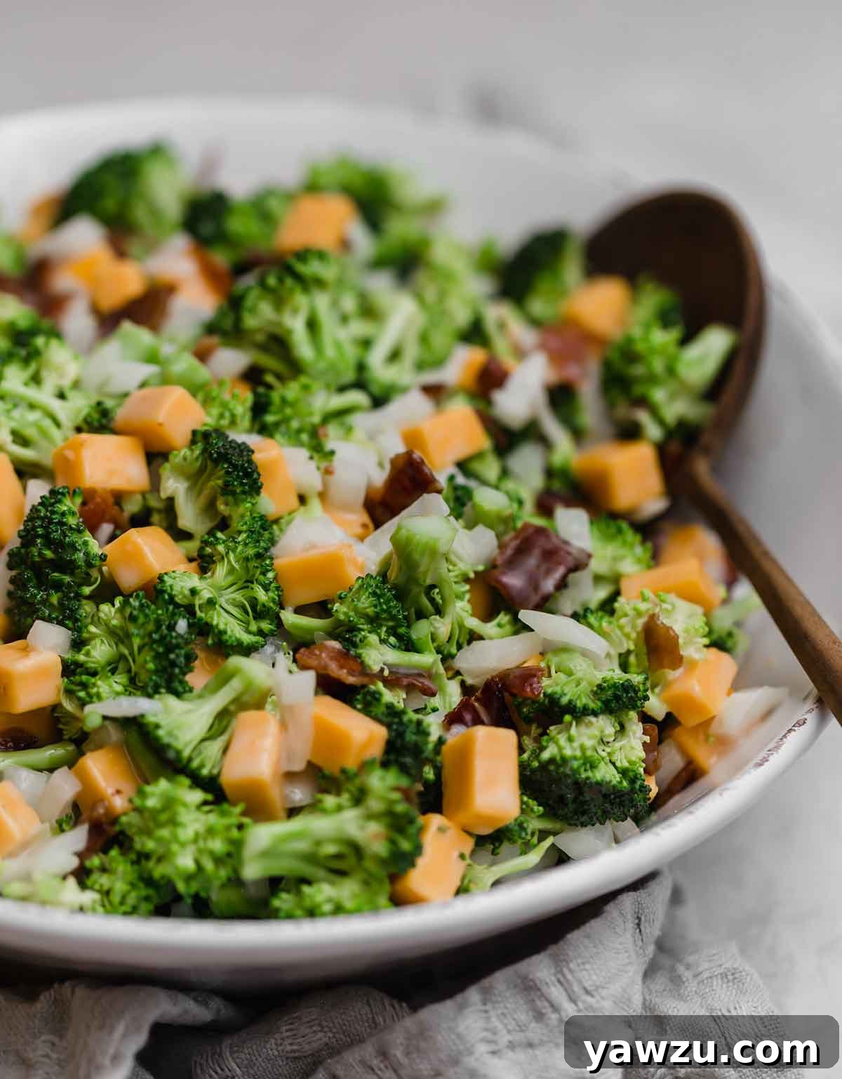 Close up photo of broccoli salad in a white bowl with a wooden serving spoon, highlighting its creamy texture and fresh ingredients.