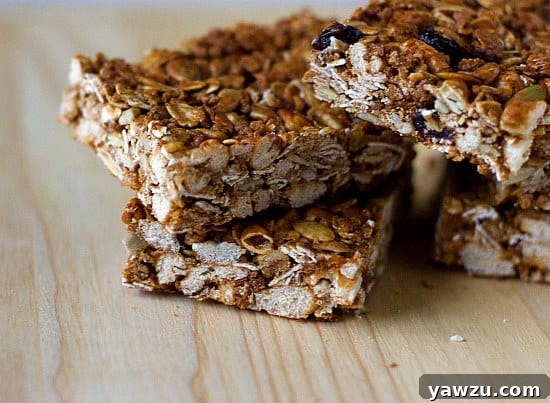 A detailed close-up of several homemade granola bars stacked together, revealing their rich texture, visible whole oats, various seeds, and colorful dried fruit pieces.