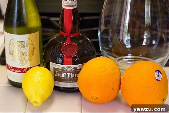 Various ingredients for white sangria laid out: a bottle of white wine, orange slices, lemon slices, a glass of orange juice, and a bottle of Grand Marnier liqueur.