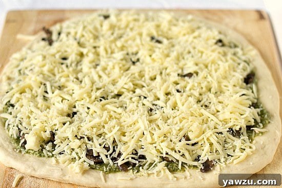 Pizza dough adorned with pesto, mushrooms, and a luxurious topping of provolone and Parmesan cheese, ready for baking.