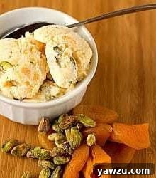Scoops of dried apricot pistachio ice cream in a white bowl with a spoon with pistachios and dried apricots next to the bowl.