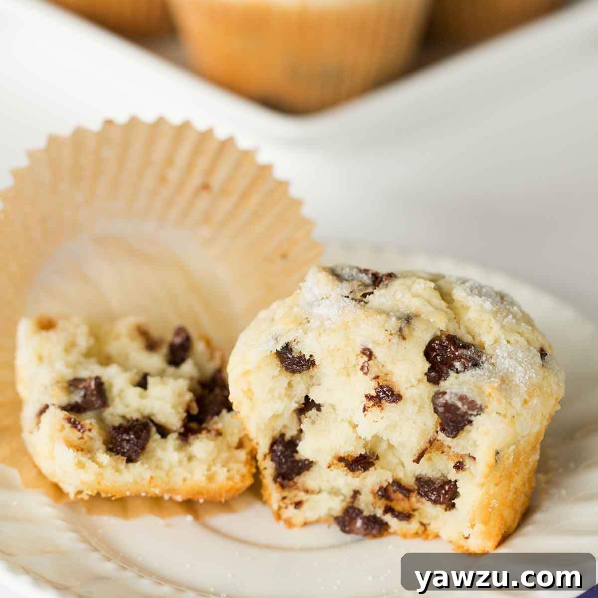 An inviting image of a chocolate chip muffin broken in half on a pristine white plate, with its paper liner resting beside it, highlighting its warm, gooey center.