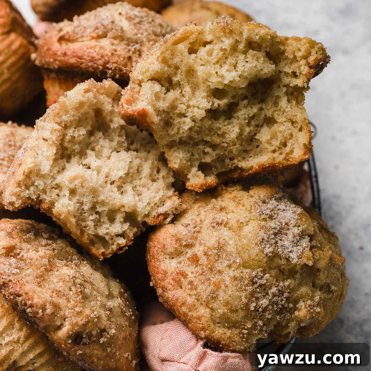 A charming basket filled with warm apple cinnamon muffins, nestled in a soft pink napkin, with one muffin artfully broken in half on top, revealing its tender interior.