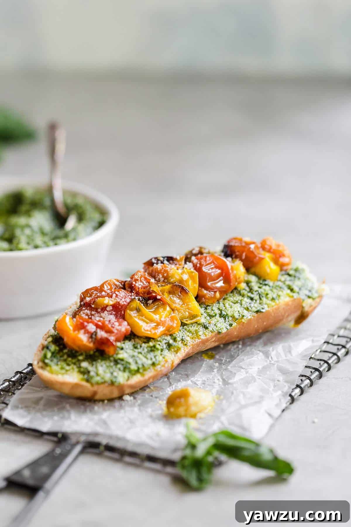 A slice of bread on a piece of parchment paper topped with pesto and roasted tomatoes with a white bowl of pesto in the back left corner.