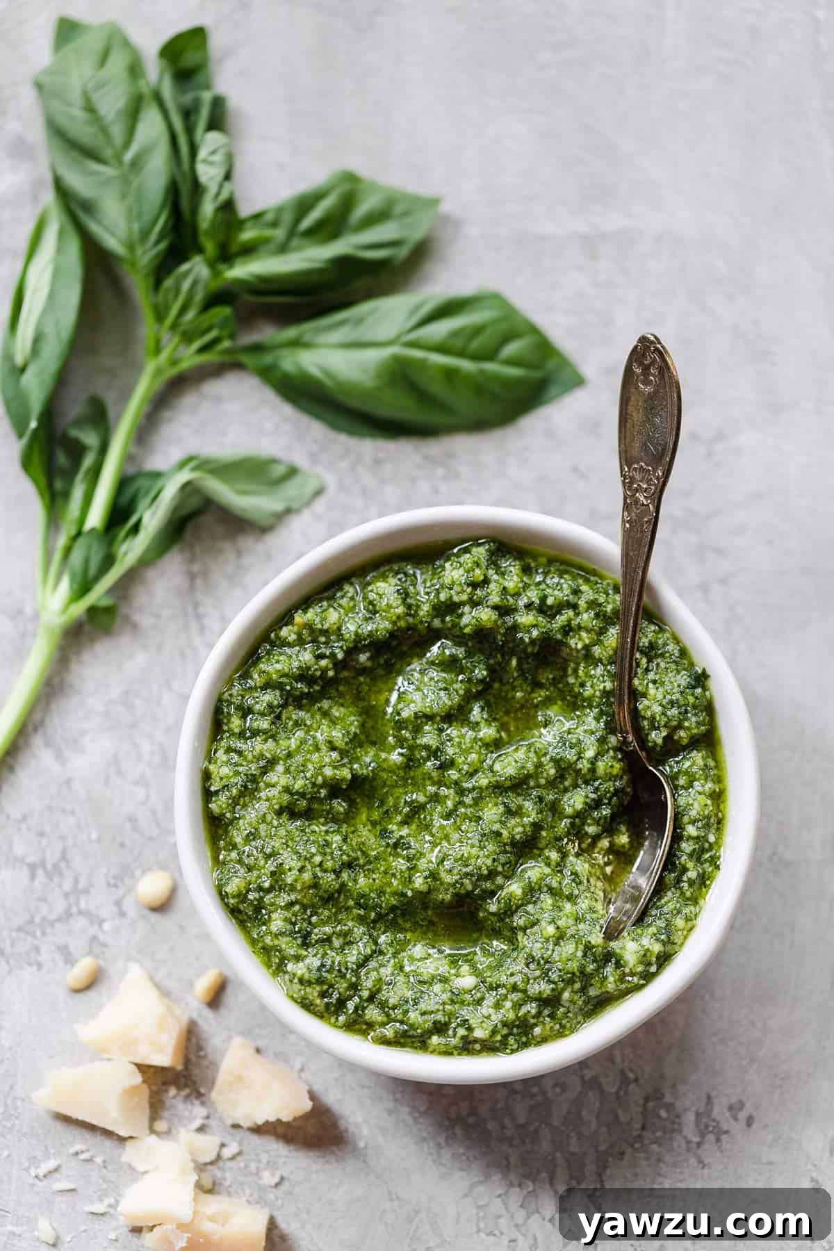 A white bowl with homemade pesto with a spoon in the right side of the bowl and fresh basil on the counter to the top left.