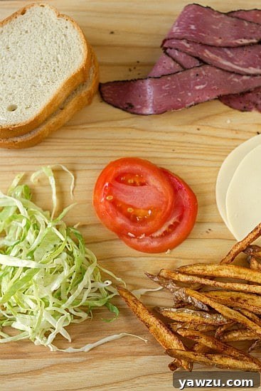 Fresh ingredients laid out for making a homemade Primanti Bros. sandwich, including sliced meats, provolone cheese, tomatoes, and containers of fries and coleslaw.
