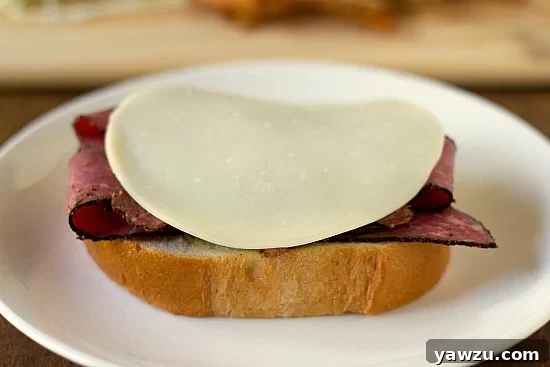 A layer of provolone cheese placed over the pastrami on the Italian bread, preparing the sandwich for the next ingredients.