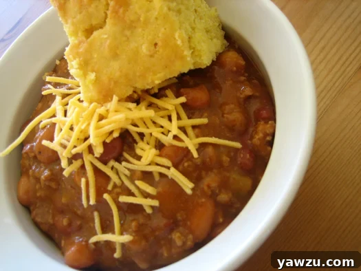 Chili in a white bowl with a piece of cornbread topped with shredded cheese.