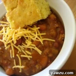 Chili in a white bowl with a piece of cornbread topped with shredded cheese.