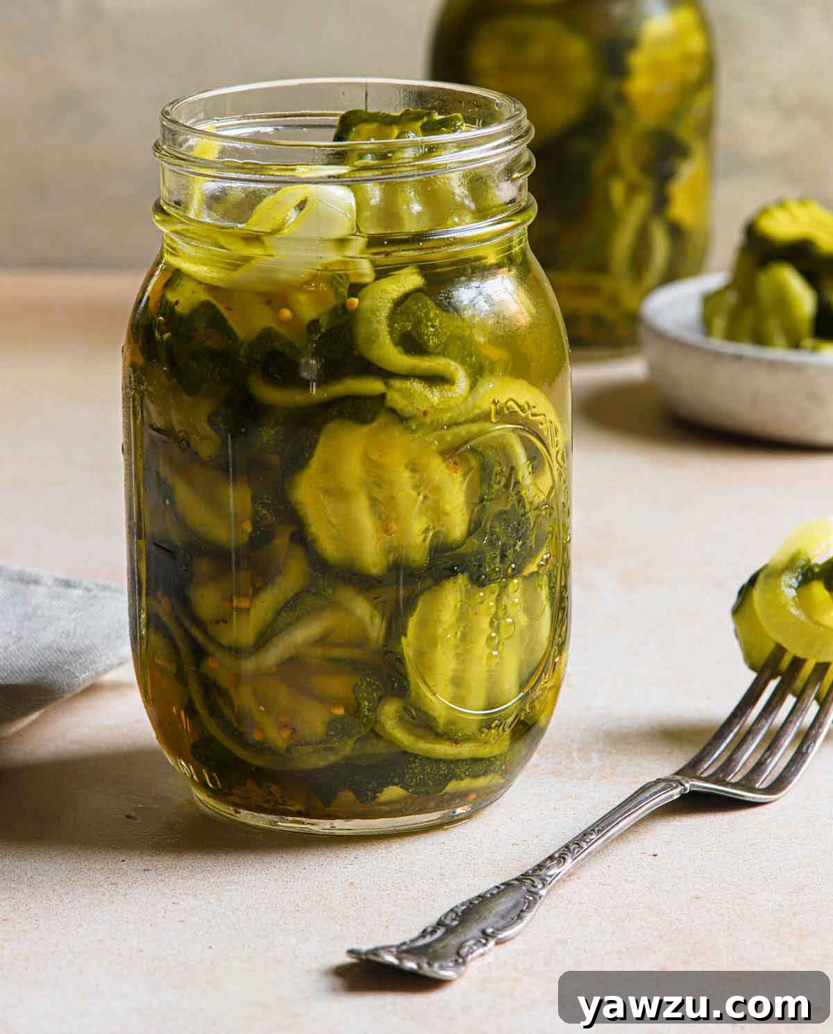 A sealed jar of homemade bread and butter pickles sits on a rustic wooden surface, with a fork holding a single, crisp pickle slice ready to be eaten.