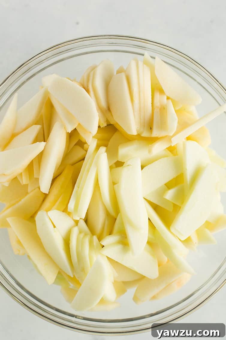 Freshly peeled, cored, and sliced McIntosh and Granny Smith apples in a clear glass bowl, glistening and ready to be mixed for the pie filling.