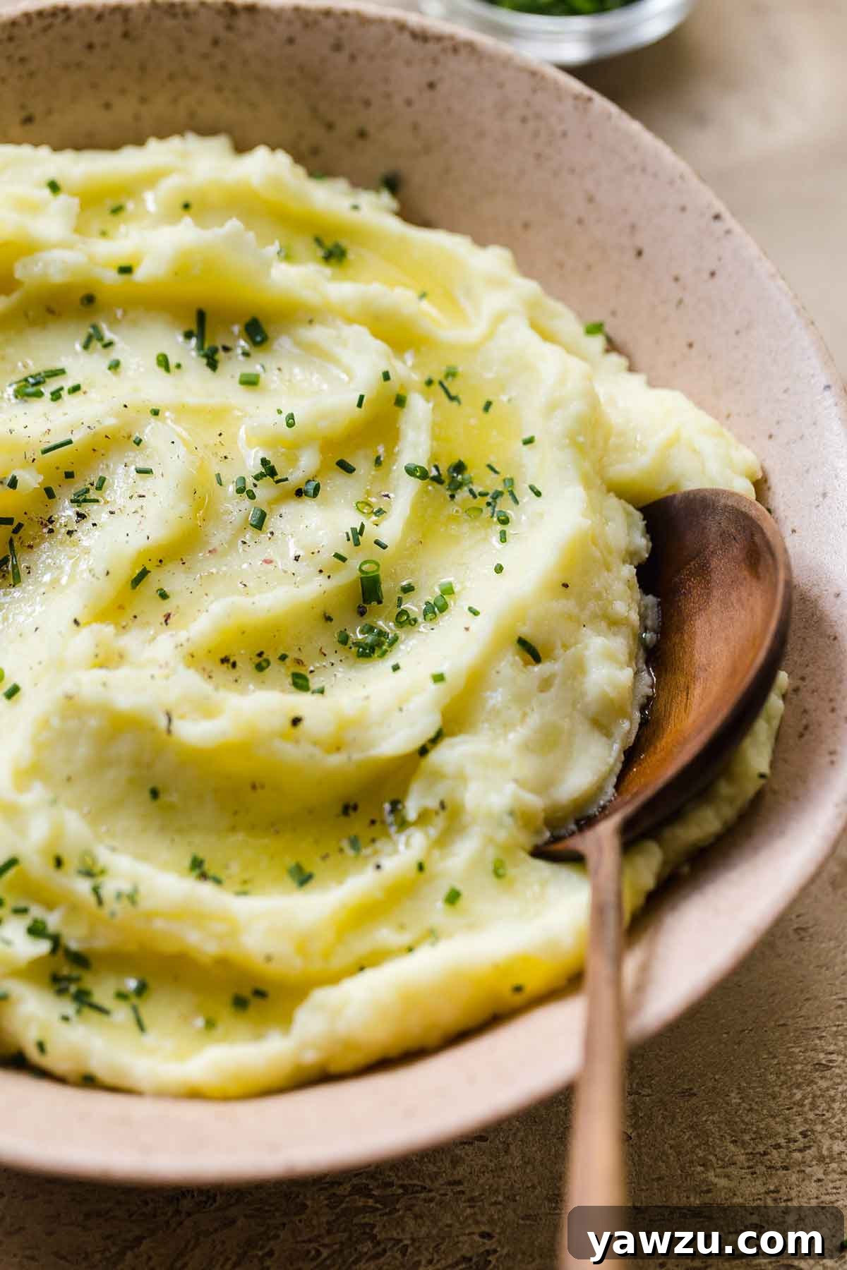 Close up photo of mashed potatoes in a serving bowl, sprinkled with minced chives, and a wooden spoon resting in the bowl.