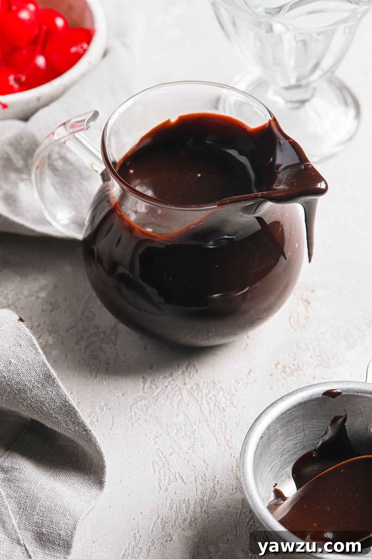 A small glass pitcher filled with hot fudge, with a bit of sauce dripping off the top, suggesting its perfect, pourable consistency.