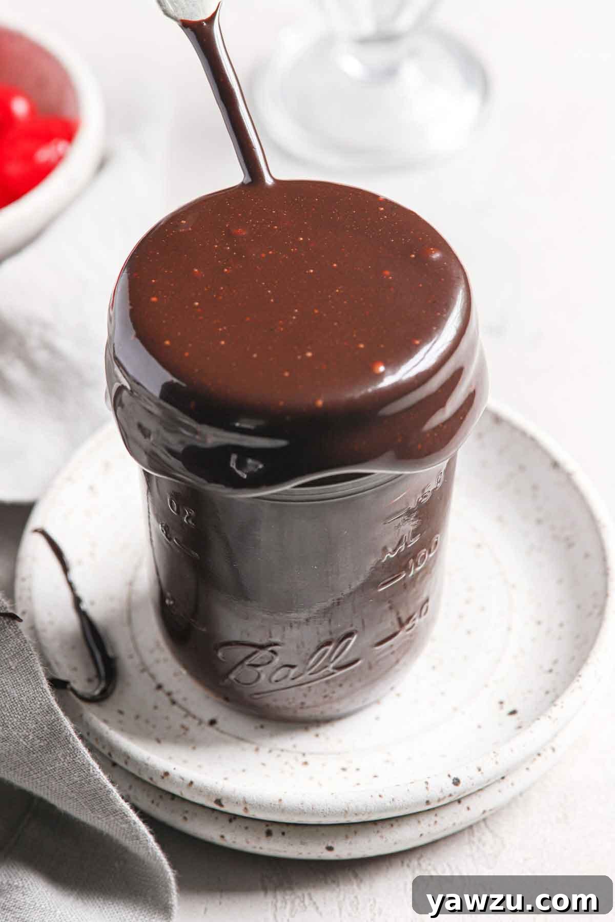 A glass jar overflowing with rich, homemade hot fudge sauce, sitting elegantly on two stacked white speckled plates.