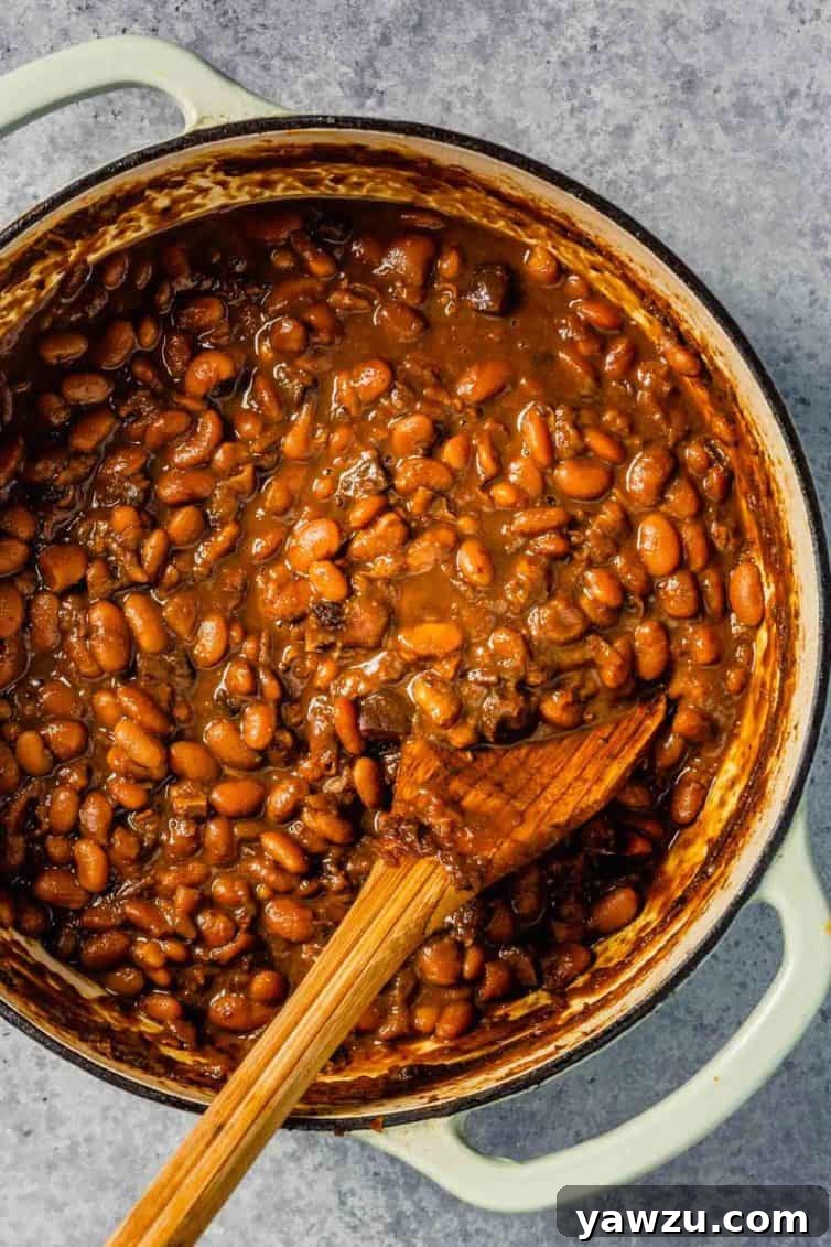 A white dutch oven filled with rich Boston baked beans, ready to be served, with a wooden spoon beside it.