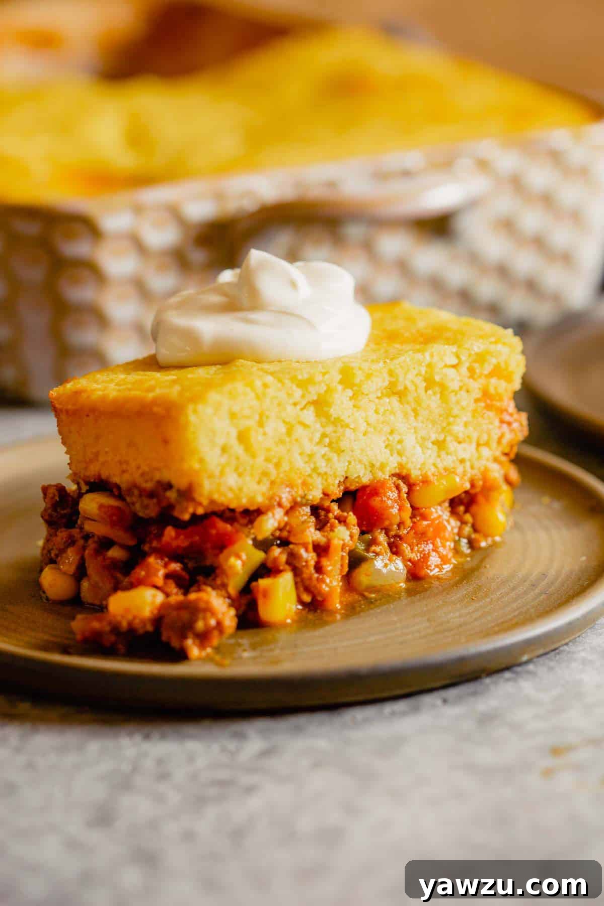 A beautifully plated serving of Tamale Pie on a brown dish, with the full casserole dish visible in the background, inviting you to dig in.