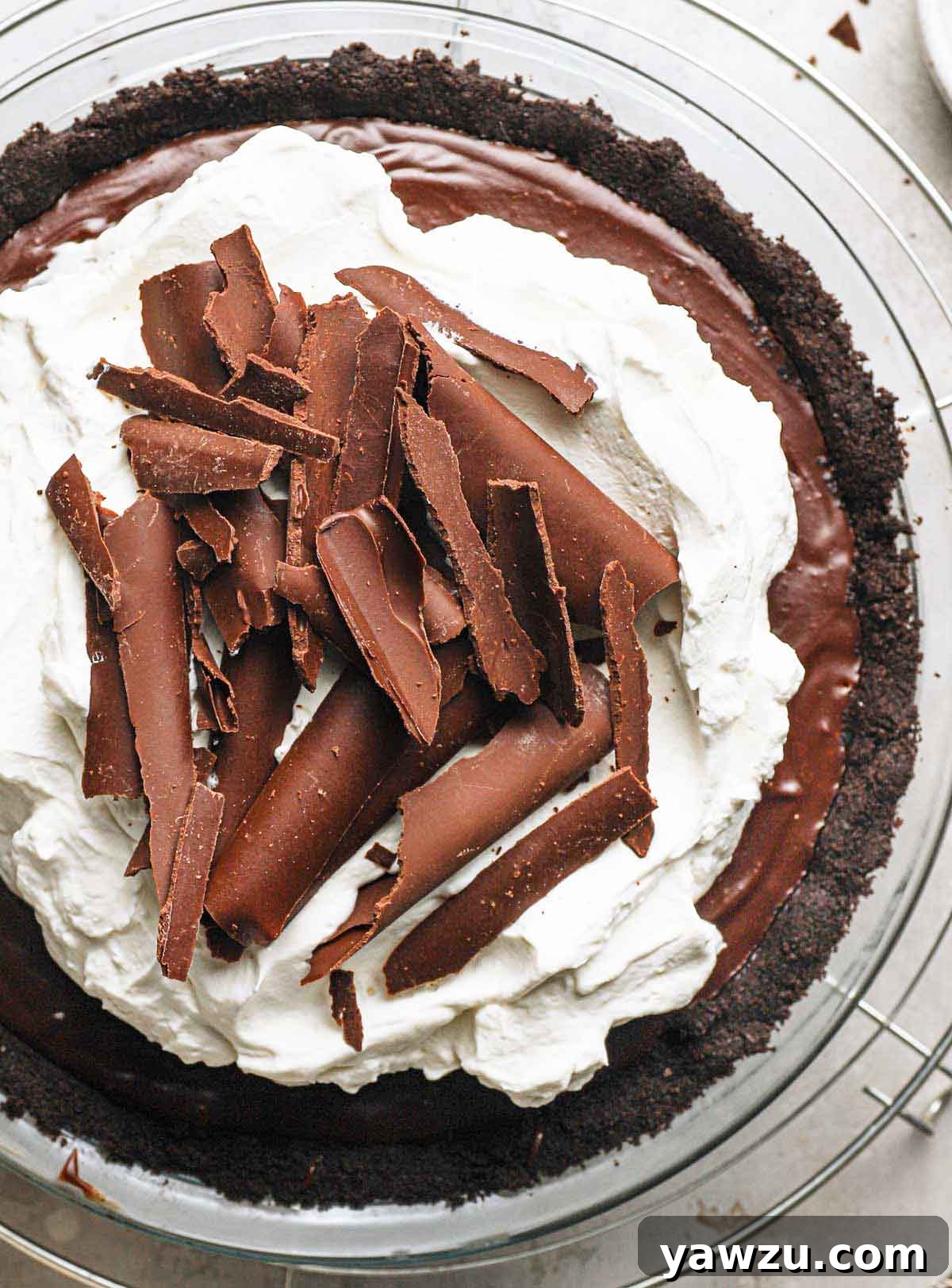 An overhead view of a whole chocolate cream pie, elegantly topped with swirling fresh whipped cream and delicate chocolate curls, presented in a clear glass pie dish.