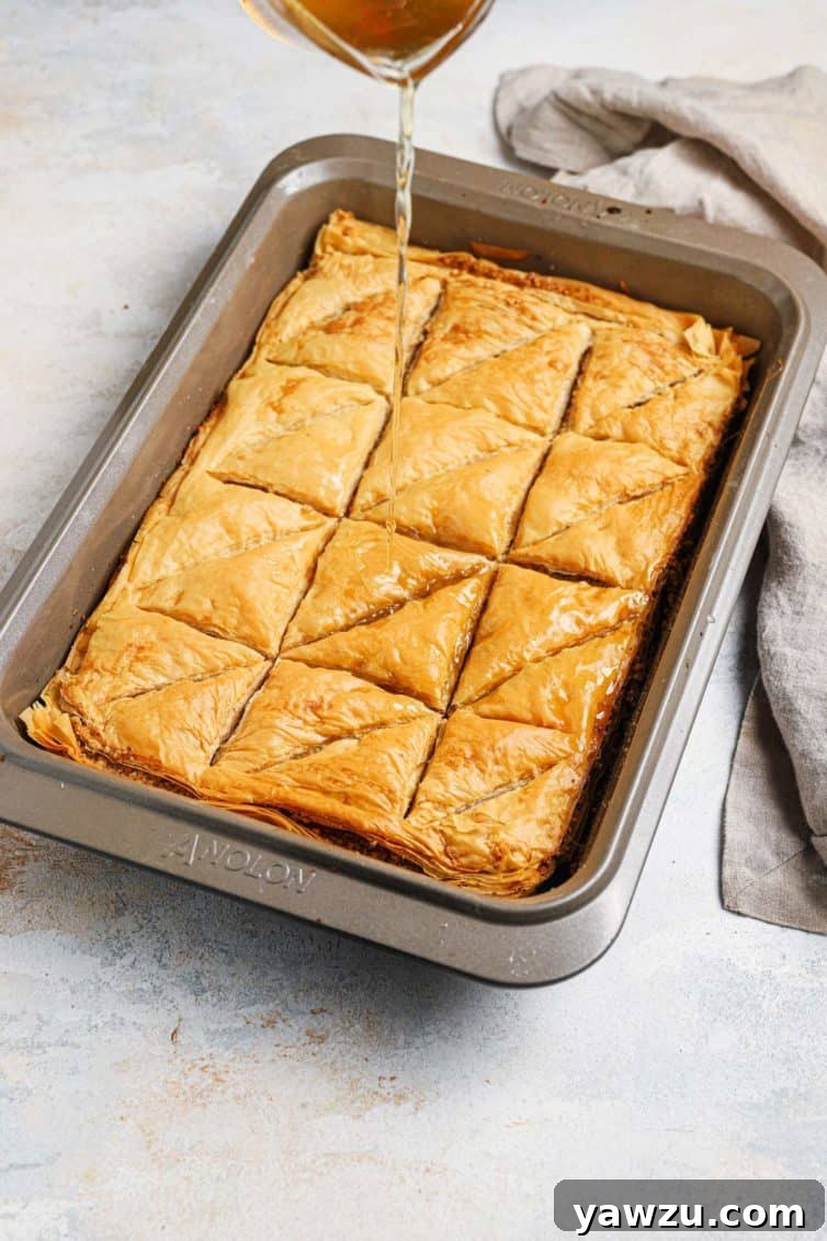 Cool syrup being slowly poured over a hot pan of freshly baked baklava, with a towel in the background.