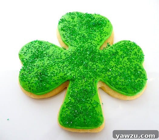 A vibrant shamrock sugar cookie, meticulously decorated with bright green royal icing and generously sprinkled with sparkling green sanding sugar, resting on a clean white surface, perfect for St. Patrick's Day.