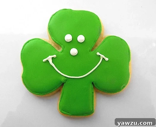 A cheerful shamrock sugar cookie, expertly coated in vivid green royal icing, featuring a distinctive white smiley face piped in the center, bringing a unique and happy twist to St. Patrick's Day treats and celebrating local tradition.