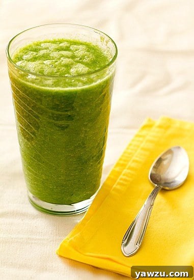 Another perspective of the green smoothie in a glass, with additional fresh fruit and green ingredients like spinach, banana, and citrus slices arranged around it, highlighting the natural components.