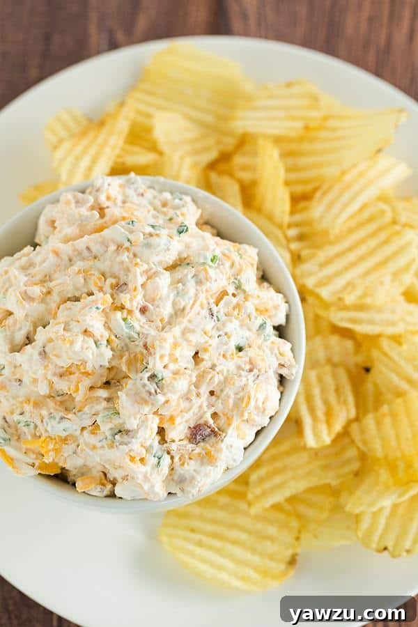 Close-up shot of the creamy Loaded Baked Potato Dip, showcasing its rich texture and visible pieces of bacon, cheese, and green onions, ready for dipping with crunchy potato chips.