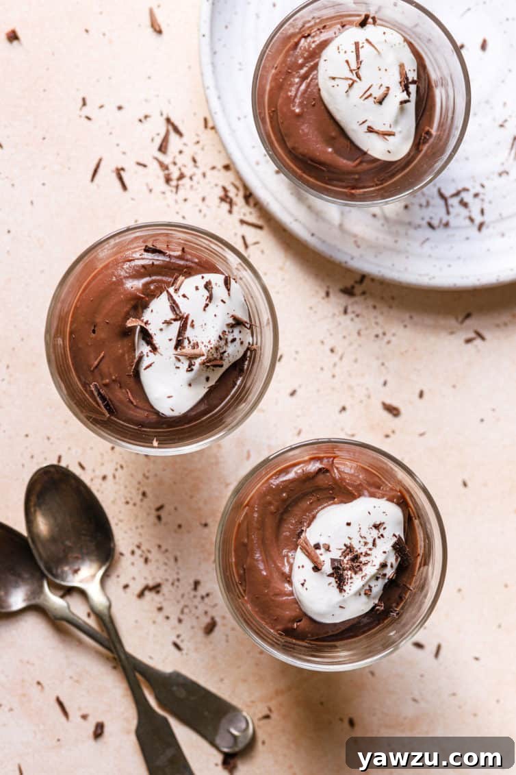 A top down photo with two glasses of pudding topped with whipped cream on the pink counter with two spoons to the left and a white plate with a glass of chocolate pudding on top.