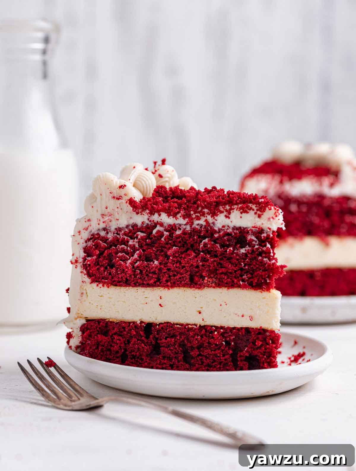 Slice of red velvet cheese on a white plate with a fork in the foreground and another slice in the background.