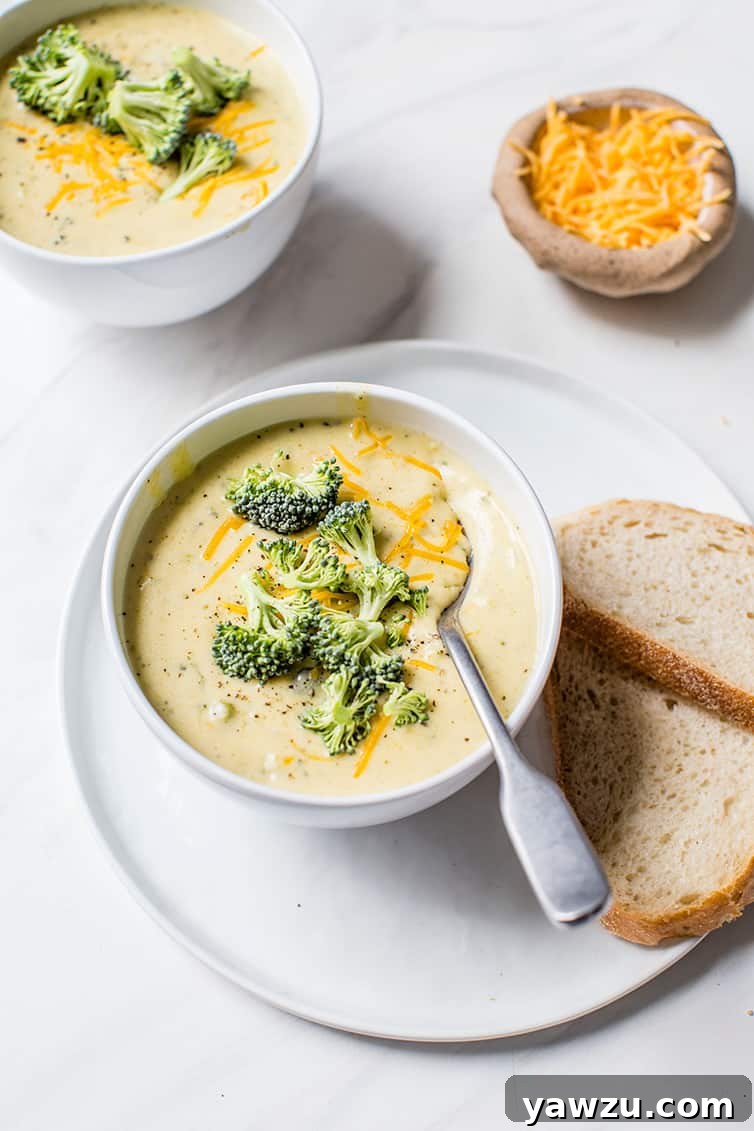 Two inviting bowls of creamy broccoli cheese soup, served on a rustic wooden table alongside slices of crusty bread and a small bowl of extra shredded cheese.