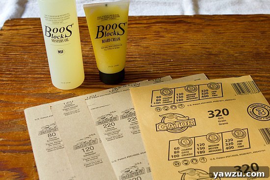Assortment of essential supplies laid out for wooden dough board restoration, including food-grade mineral oil, beeswax cream, and various grit sandpapers