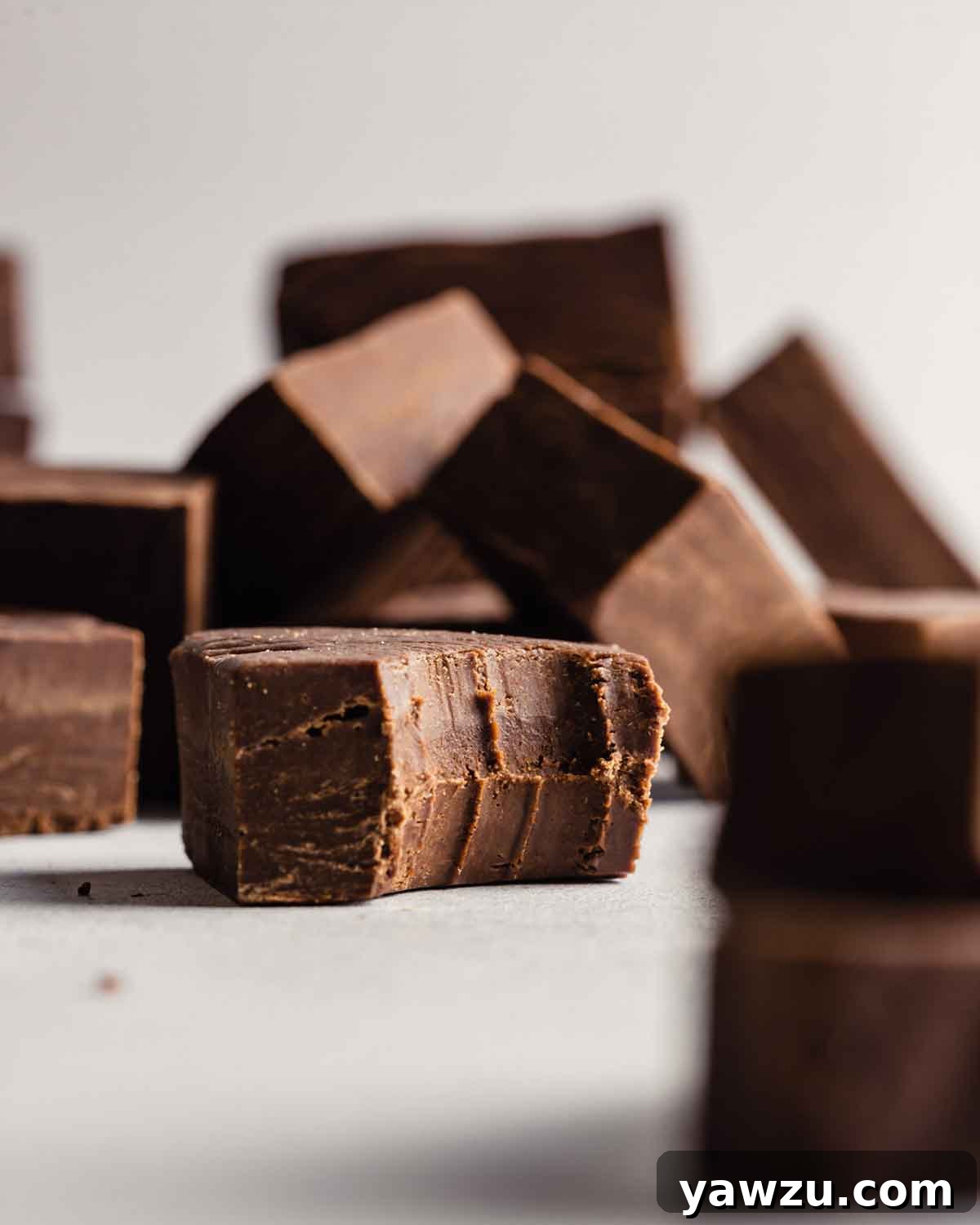 Decadent, creamy chocolate fudge square with a bite taken out, resting next to a stack of perfectly cut fudge pieces.