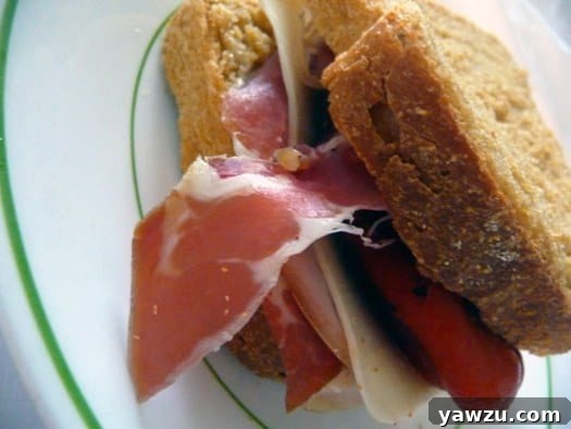 A delectable sandwich crafted with Anadama bread, generously filled with prosciutto, fresh mozzarella, and vibrant roasted red peppers, presented on a white plate.