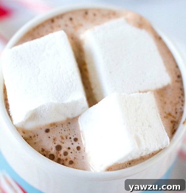 A mug of hot chocolate adorned with two homemade peppermint marshmallows, with a few extra marshmallows scattered around.