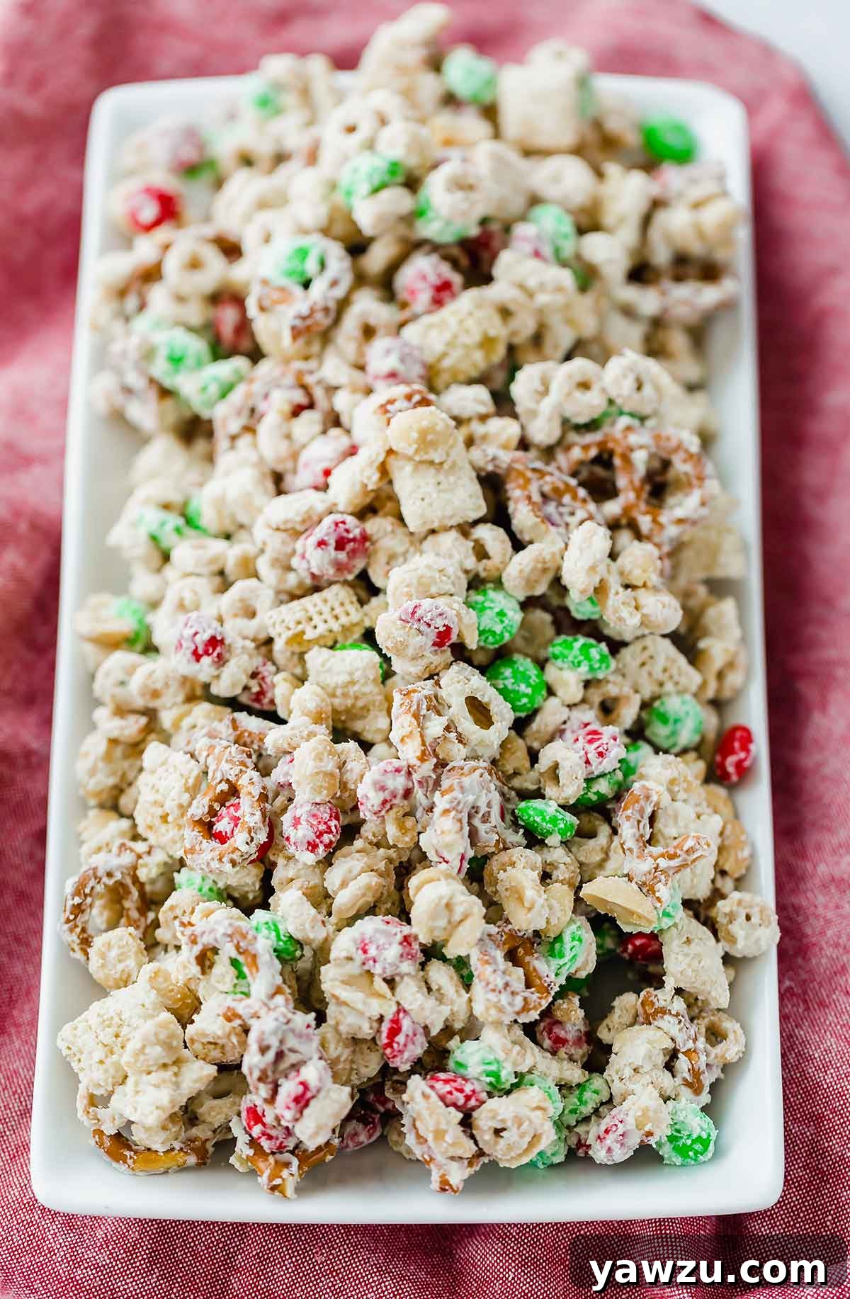 A white tray set on a red background with festive reindeer chow snack mix on it, adorned with red and green M&Ms.